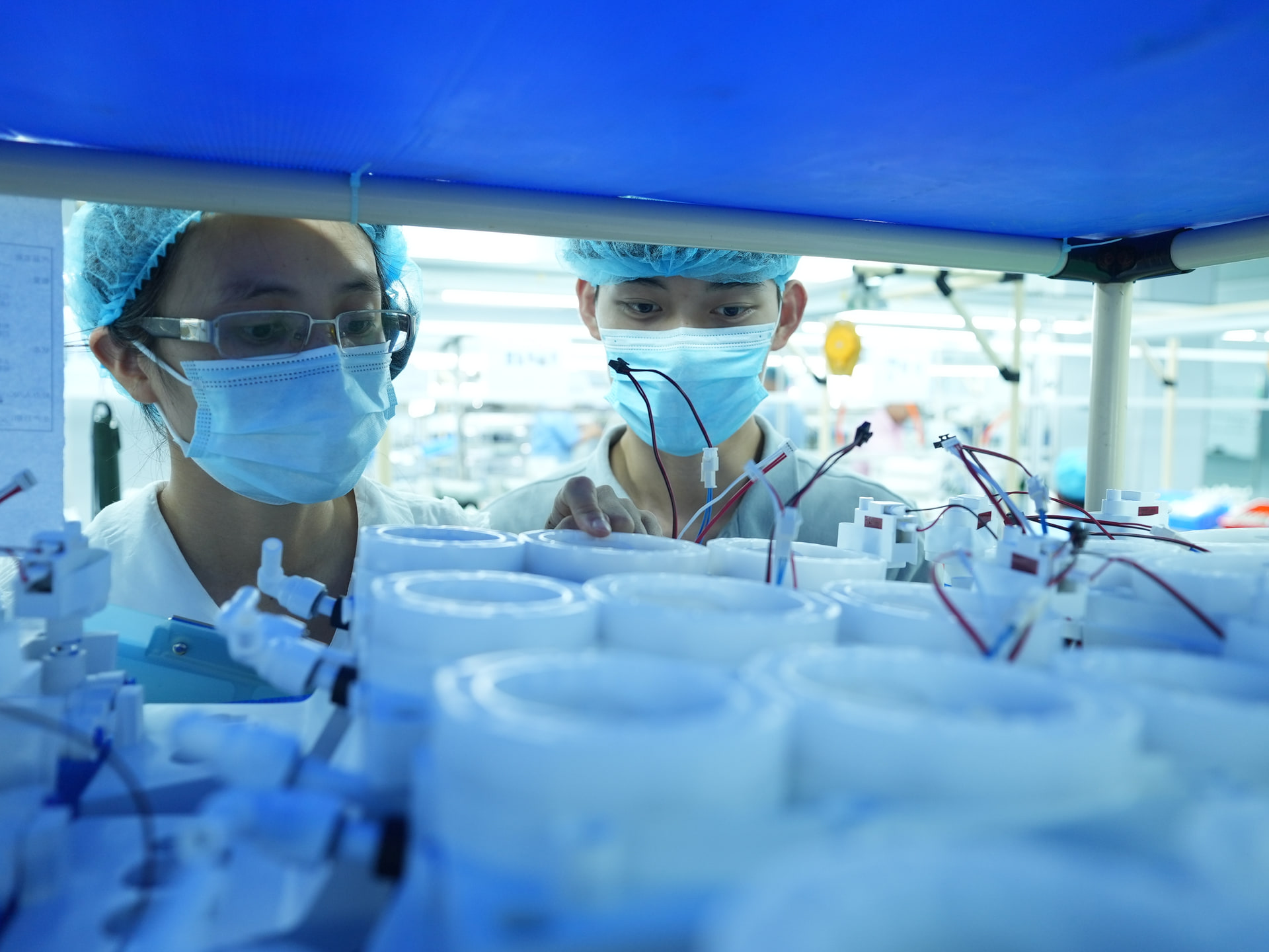 Two masked production workers inspecting assembled components under a workbench canopy.